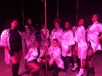 Group of women in pink satin robes posing around a dance pole in a dimly lit pole dance studio with magenta club lighting