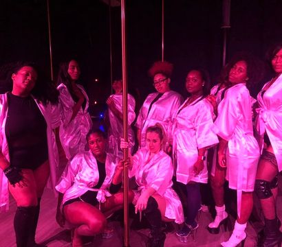 Group of women in pink satin robes posing around a dance pole in a dimly lit pole dance studio with magenta club lighting