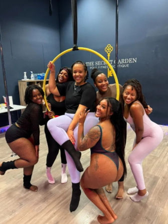 Group of six women smiling and posing around a yellow aerial hoop (lyra) in an indoor pole fitness studio, wearing activewear and tights during a dance/aerial class.