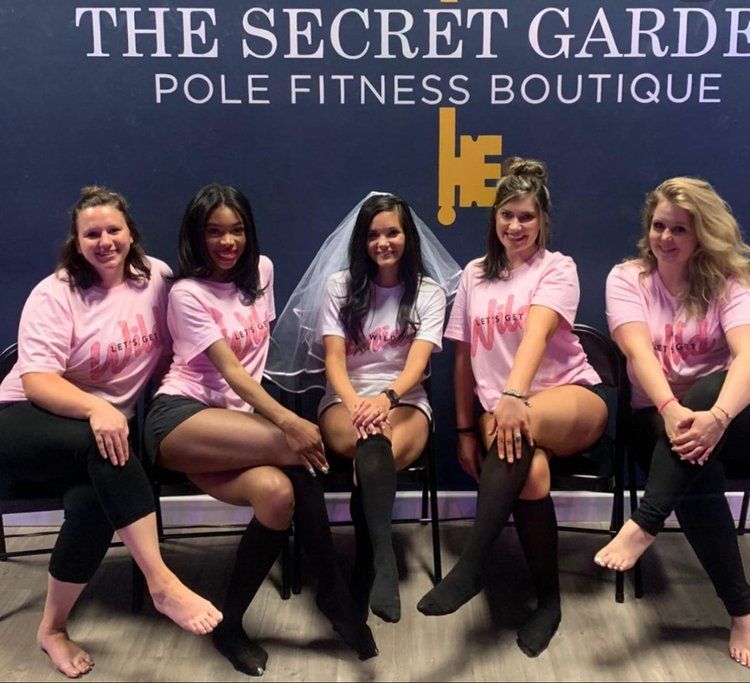 Five women in matching pink shirts, the center wearing a bridal veil, seated and posing at a pole fitness studio for a bachelorette party.