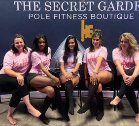 Five women in matching pink shirts, the center wearing a bridal veil, seated and posing at a pole fitness studio for a bachelorette party.