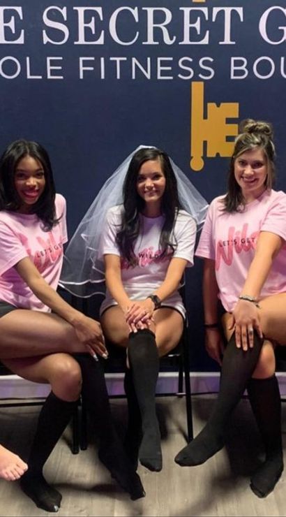 Five women in matching pink shirts, the center wearing a bridal veil, seated and posing at a pole fitness studio for a bachelorette party.