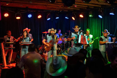 Live norteño/regional Mexican band on an indoor stage with accordion, guitar, drums, keyboard and congas; musicians in cowboy hats and denim vests under colorful stage lights, audience in foreground.