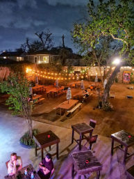 Nighttime outdoor beer-garden patio with wooden picnic tables and striped umbrellas, warm string lights strung between trees and a fence, a colorful mural on the fence, and a few patrons at high-top tables in an urban backyard setting.