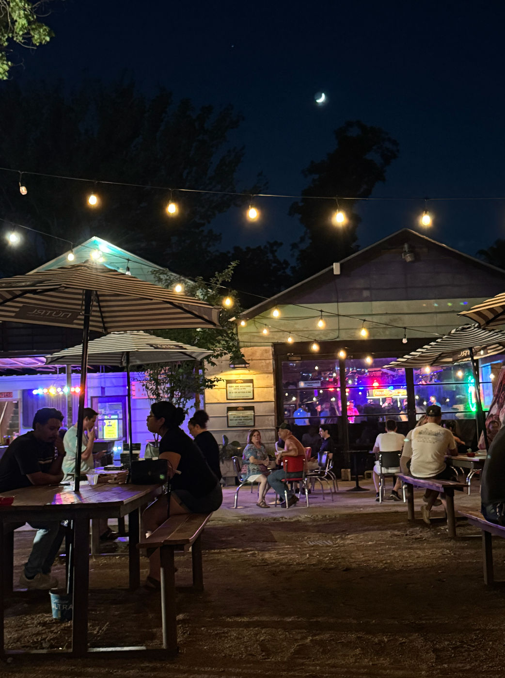 Nighttime outdoor bar patio with hanging string lights and striped umbrellas, groups seated at picnic tables, colorful neon-lit interior with live-music glow behind glass, and a crescent moon overhead.