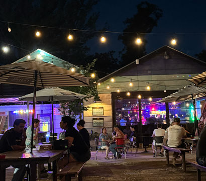 Nighttime outdoor bar patio with hanging string lights and striped umbrellas, groups seated at picnic tables, colorful neon-lit interior with live-music glow behind glass, and a crescent moon overhead.