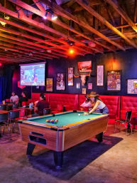 Neon-lit sports bar interior with exposed wooden beams, red tufted banquette, framed posters and steer skull, patrons watching a projected game while a woman in a hat plays pool.