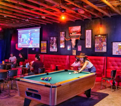 Neon-lit sports bar interior with exposed wooden beams, red tufted banquette, framed posters and steer skull, patrons watching a projected game while a woman in a hat plays pool.