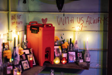 Cozy indoor memorial shrine on a wooden shelf with warm candlelight, framed photos, marigold flowers, glass bottles and a bright red container beneath handwritten wall text 'WITH US ALWAYS' and heart doodles.