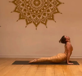 Yoga practitioner in upward-facing dog pose on a black mat inside a warm yoga studio with a large mandala wall mural and hardwood floor.