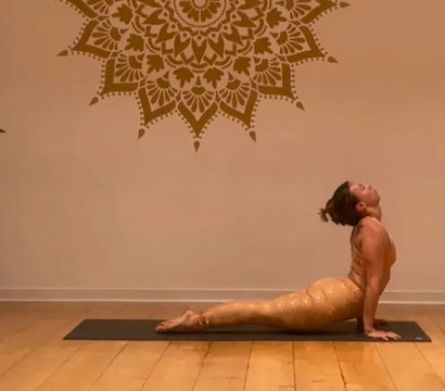 Yoga practitioner in upward-facing dog pose on a black mat inside a warm yoga studio with a large mandala wall mural and hardwood floor.