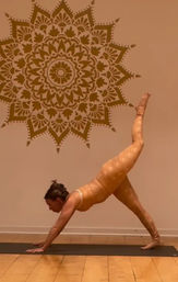 Yoga practitioner holding a three-legged downward dog on a black mat in a warm-toned indoor studio with hardwood floor and a large decorative mandala wall mural.