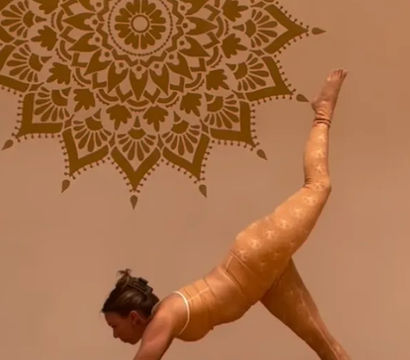 Yoga practitioner holding a three-legged downward dog on a black mat in a warm-toned indoor studio with hardwood floor and a large decorative mandala wall mural.