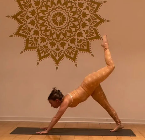 Person in orange activewear in three‑legged downward dog on a black yoga mat inside a warm-toned studio with wooden floor and a large mandala wall mural.