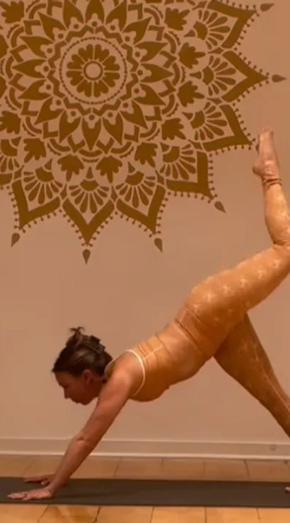 Person in orange activewear in three‑legged downward dog on a black yoga mat inside a warm-toned studio with wooden floor and a large mandala wall mural.