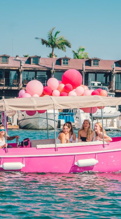 Pink electric party boat with a canopy and pink balloons carrying laughing women through a sunny marina harbor