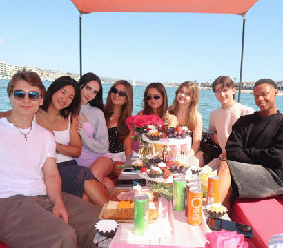 Group of eight young adults enjoying a sunny boat party in a coastal harbor, seated on a pink boat around a picnic table with roses, cupcakes, fruit and canned drinks.