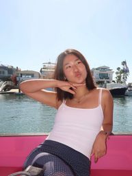 Playful woman posing on a pink boat seat at a sunny coastal marina, with yachts and waterfront homes lining the harbor