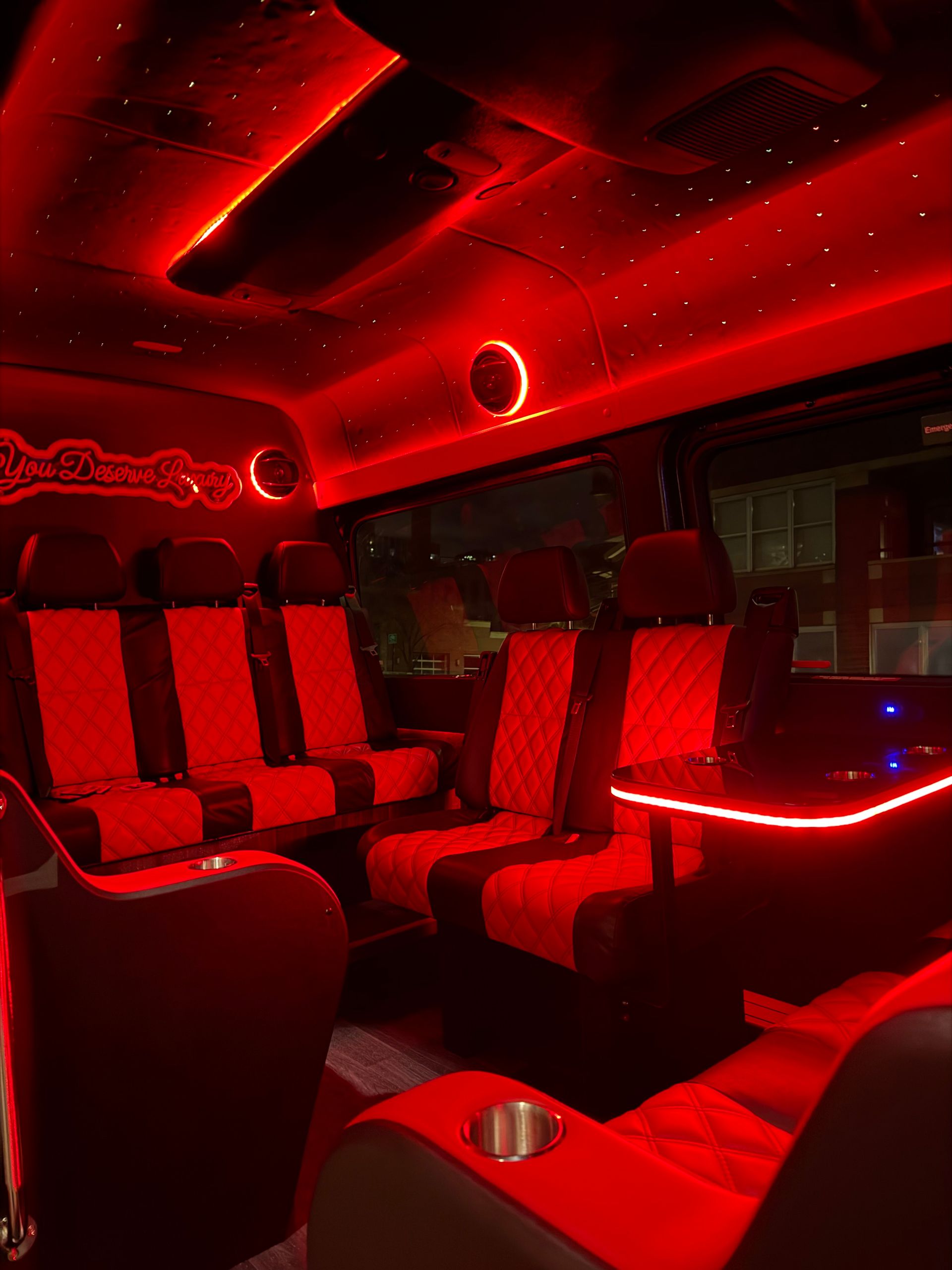 Luxury party van interior with red LED ambient lighting, diamond-quilted leather seats, illuminated side table and cup holders, and a starry fiber-optic ceiling.