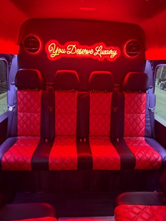 Red-lit luxury passenger van interior with quilted red-and-black leather bench seats, starry ceiling lights and a glowing neon sign reading "You Deserve Luxury".