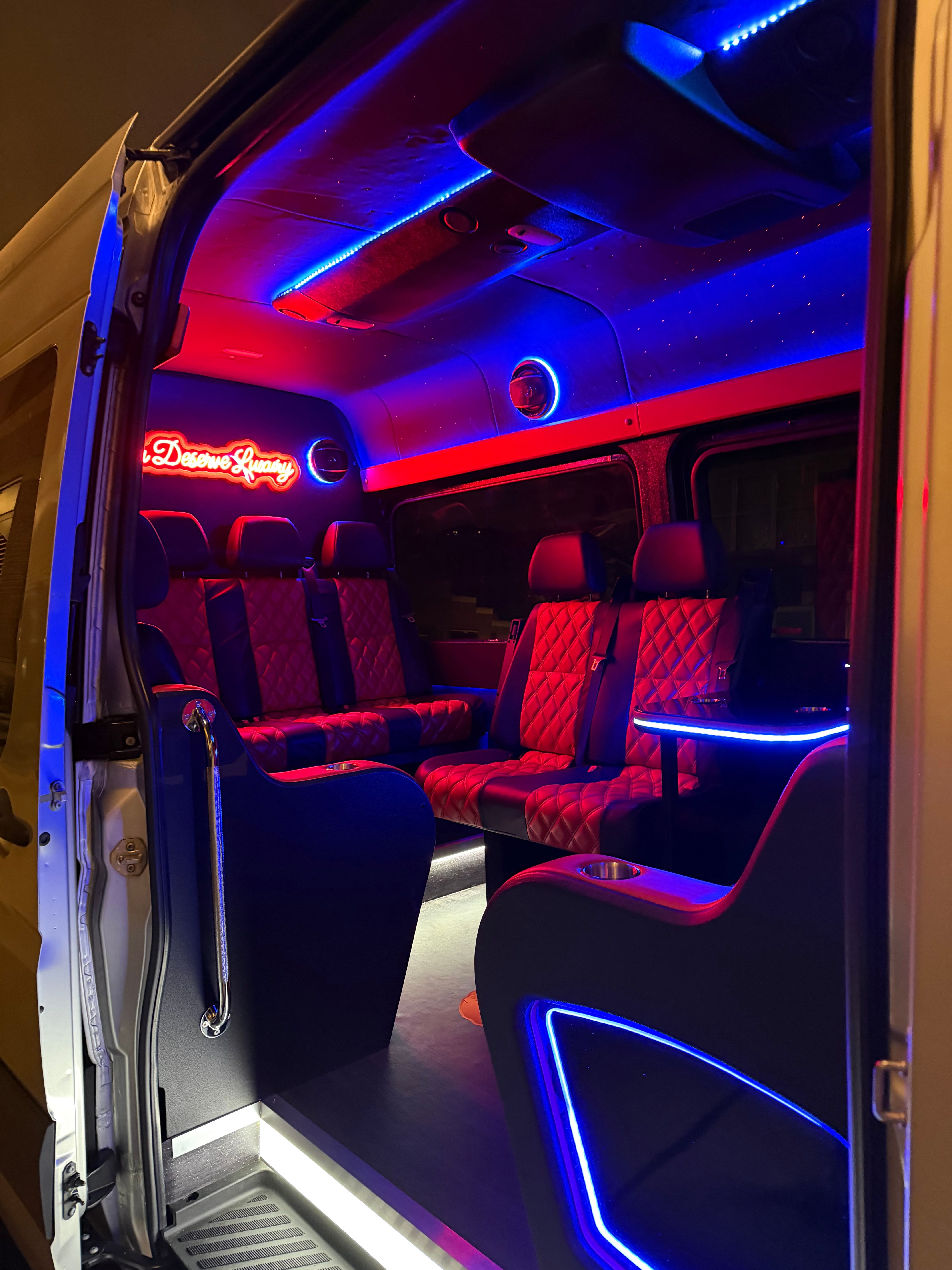 Vibrant luxury shuttle van interior at night with red quilted leather seats, blue and red LED neon accents, illuminated side table and cup holders, and an open sliding door.
