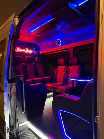 Vibrant luxury shuttle van interior at night with red quilted leather seats, blue and red LED neon accents, illuminated side table and cup holders, and an open sliding door.