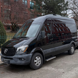 Sleek black Mercedes Sprinter high‑roof van parked on a downtown street, glossy side reflecting red brick buildings and bare trees on an overcast day.