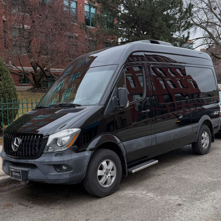 Sleek black Mercedes Sprinter high‑roof van parked on a downtown street, glossy side reflecting red brick buildings and bare trees on an overcast day.
