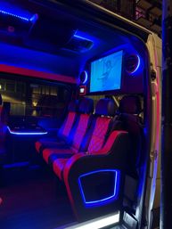 Luxurious party van interior at night with red quilted leather seats, vibrant blue LED accent lighting, built-in screen showing an aerial performer, cupholders and city buildings visible outside.