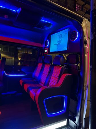 Luxurious party van interior at night with red quilted leather seats, vibrant blue LED accent lighting, built-in screen showing an aerial performer, cupholders and city buildings visible outside.