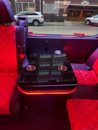Luxury shuttle interior with red quilted leather seats, glossy black fold-down table with four cup holders and red LED accent lighting, window revealing parked cars on a city street outside