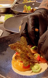 Gloved chef plating a colorful heirloom tomato and microgreen salad on a black ceramic plate in a busy restaurant kitchen.