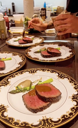 Chef’s hands saucing medium-rare beef tenderloin with asparagus and gravy on ornate gold-rimmed plates in a fine-dining kitchen — gourmet plating for a restaurant meal.
