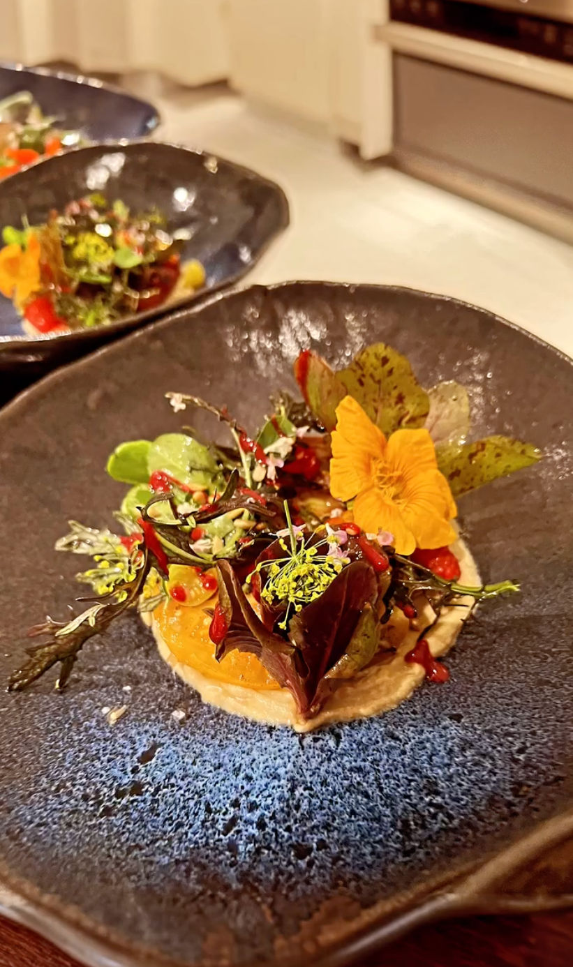 Fine-dining plated salad: microgreens, mixed greens and a bright orange edible flower atop a creamy puree with red vinaigrette dots in a textured blue ceramic bowl