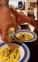 Hands drizzling olive oil over Italian-style filled pasta in a creamy yellow sauce, garnished with herbs in blue-rimmed bowls on a kitchen counter.