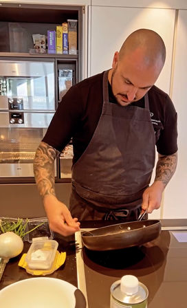 Bald tattooed cook in a dark apron focused on stirring a sauté pan on a modern kitchen stovetop, with onion, green beans and seasoning containers nearby.