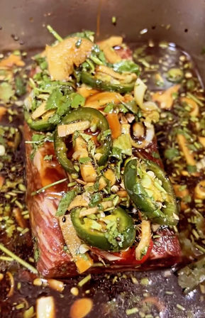 Raw marinated beef slab in a shallow pan, glazed in a dark soy-style sauce and topped with sliced jalapeños, cilantro, garlic and ginger — zesty, Asian-inspired prep ready to cook.