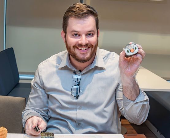 Smiling man in a light button-up shirt with glasses on his collar, wearing disposable gloves and holding a cute penguin-faced sushi roll during a hands-on sushi-making class in an indoor workshop.