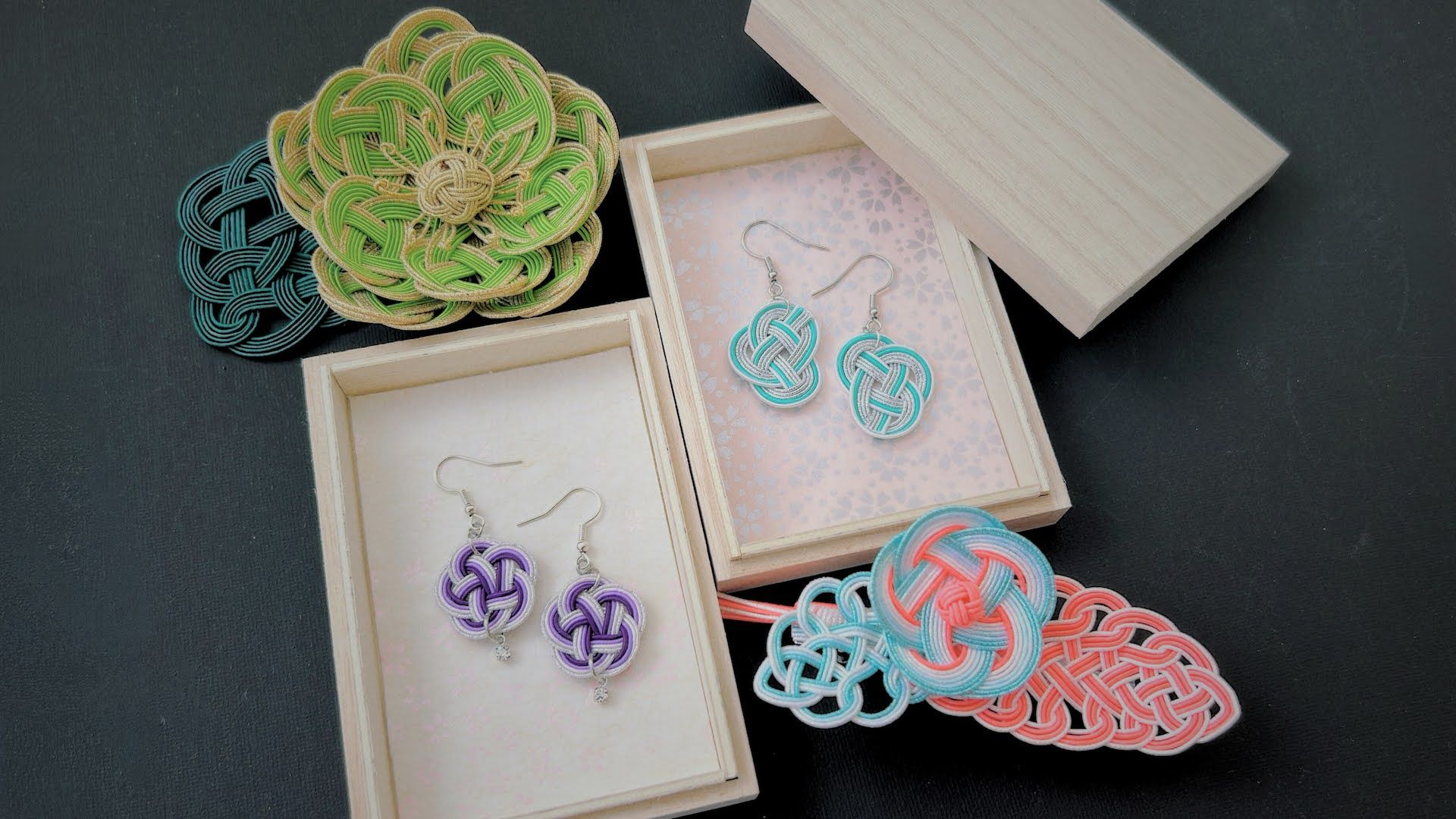 Colorful handmade mizuhiki knot earrings and braided cord accessories in pastel teal, coral, purple and green displayed in wooden gift boxes on a dark background