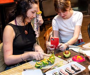 Two friends at an indoor sushi-making workshop crafting avocado sushi rolls on a wooden table with gloves, rolling mat, chopsticks and a smartphone.