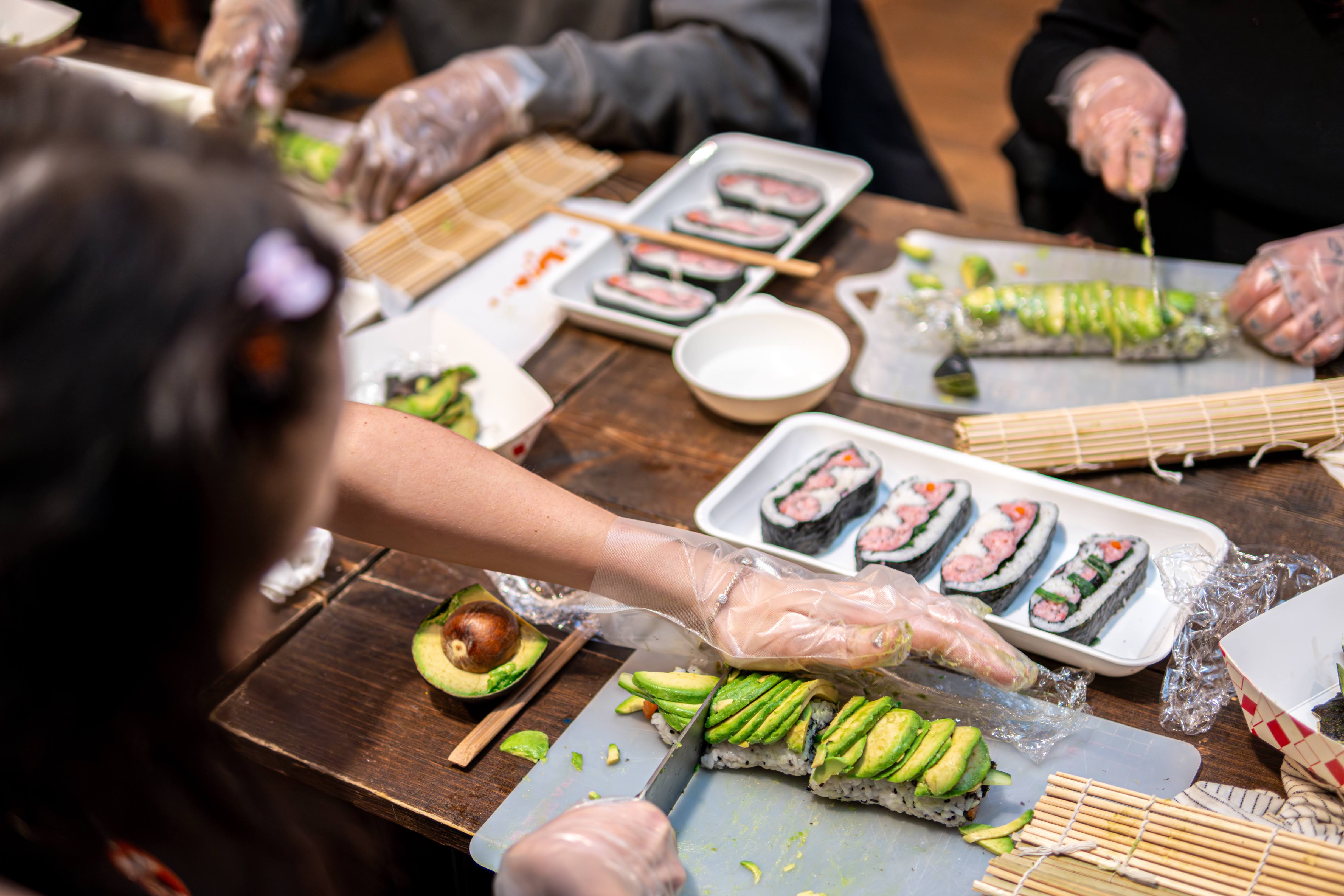 Hands-on sushi-making workshop: gloved hands slice avocado-topped sushi rolls on a wooden table with bamboo mats, chopsticks, avocado halves and plated sushi pieces.