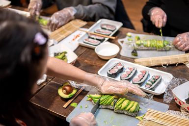 Hands-on sushi-making workshop: gloved hands slice avocado-topped sushi rolls on a wooden table with bamboo mats, chopsticks, avocado halves and plated sushi pieces.