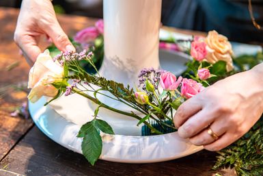 Close-up of hands arranging pink and peach roses, filler blooms and greenery around a white vase on a wooden table to create a DIY floral arrangement centerpiece