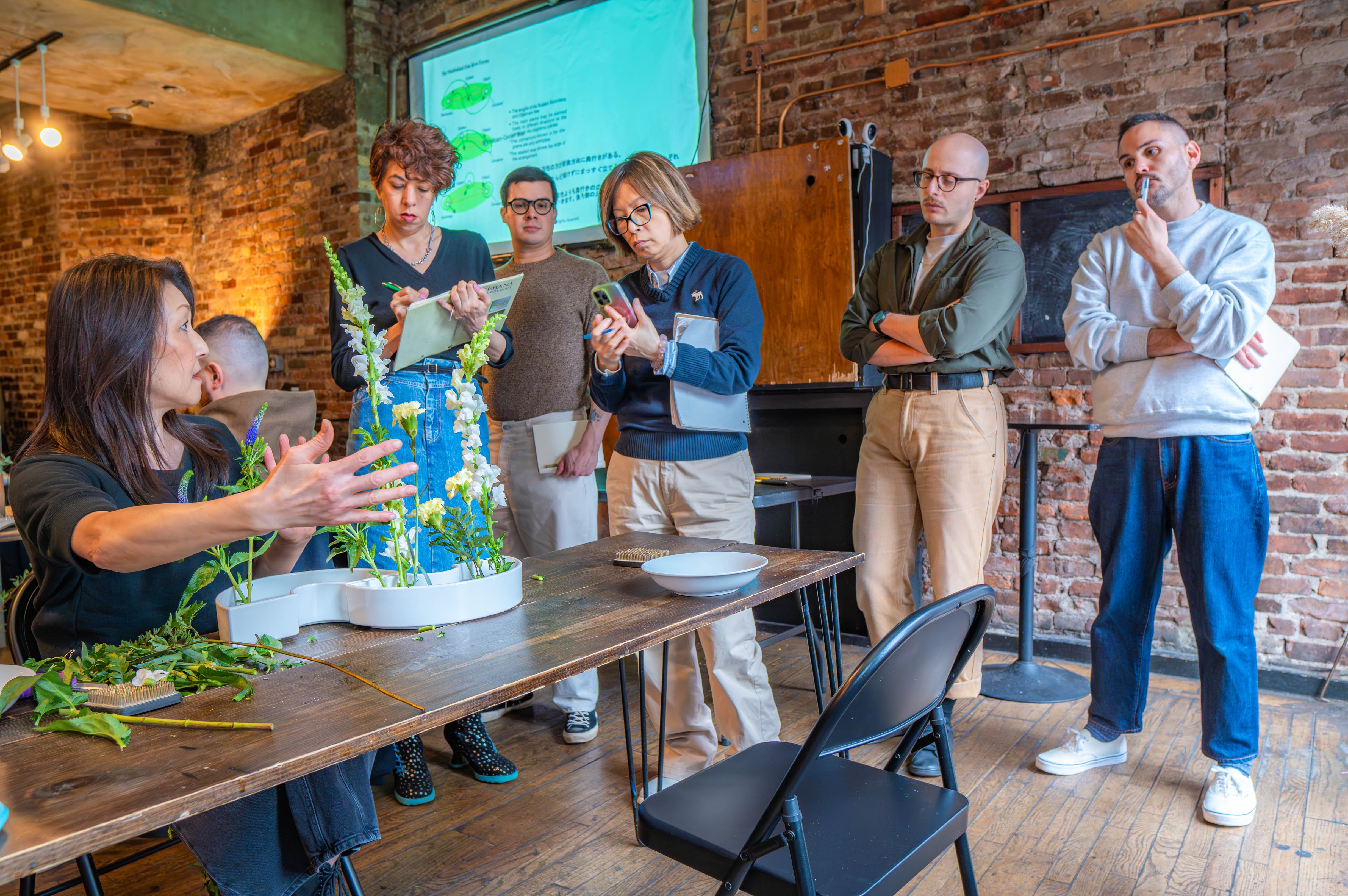 Hands-on flower-arranging workshop in a cozy urban loft: instructor demonstrating white tray arrangements on a wooden table while a small group watches, takes notes and photographs against exposed brick walls and wooden floors.