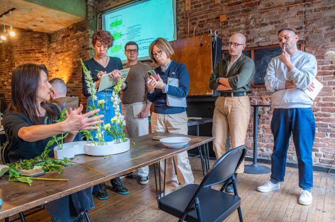 Hands-on flower-arranging workshop in a cozy urban loft: instructor demonstrating white tray arrangements on a wooden table while a small group watches, takes notes and photographs against exposed brick walls and wooden floors.