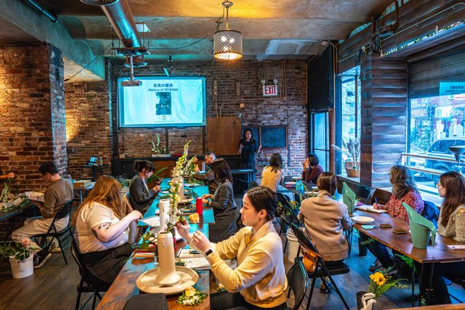 Ikebana flower-arranging workshop in a cozy brick-walled urban studio, communal tables of students arranging stems and tools while an instructor presents "History of IKEBANA" on a projector screen.