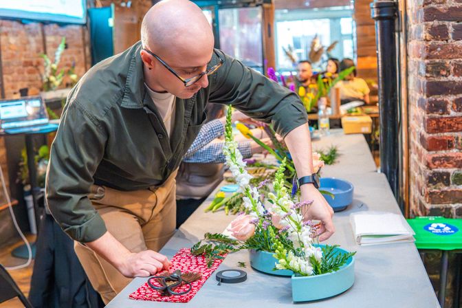 Person arranging pink roses, white snapdragons and greenery in a blue semicircular vase during a hands-on floral arrangement workshop at a rustic brick-walled studio, with scissors and floral tape on the table.