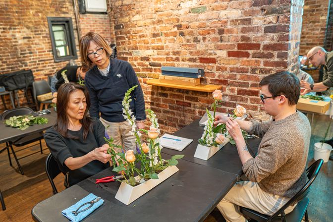 Floral arranging workshop in a brick-walled studio with participants arranging peach roses, white snapdragons and greenery in long white containers.