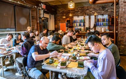 Lively sushi-making class in a cozy brick-walled urban studio, adults seated at long tables rolling nori and rice on bamboo mats under pendant lights — hands-on cooking workshop and team-building activity.