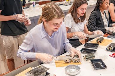 Hands-on sushi-making class at a kitchen counter with participants wearing gloves slicing nori-wrapped rolls on bamboo mats into pieces
